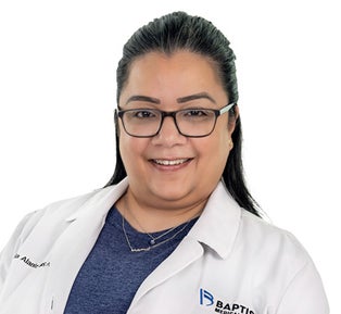 Professional headshot of a healthcare provider wearing a white medical coat and dark-framed glasses against a light gray background. The individual is facing the camera with a direct gaze. Visible branding is embroidered on the coat, including a healthcare organization logo. The image is well-lit, high-resolution and suitable for use in provider directories, website profiles, marketing materials and professional publications.