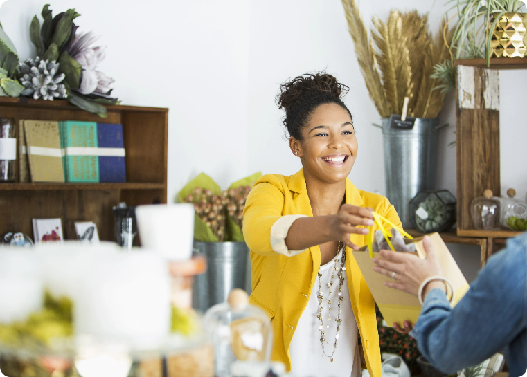 Lady in a retail shop behind the counter handing a customer their package and smiling
