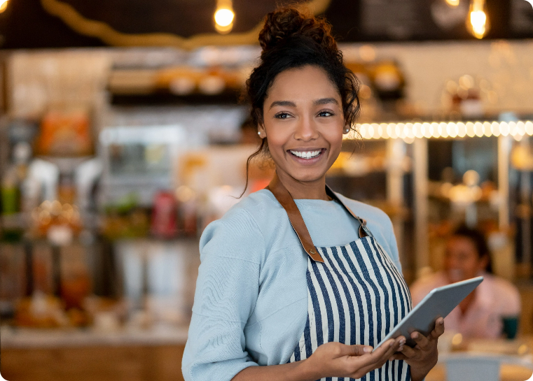 Lady in a striped apron, holding a tablet standing in a business while smiling out at the customers