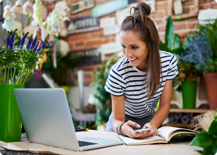 Lady leaning over a table, with flowers and a computer on it, typing on her phone while reading something off the computer