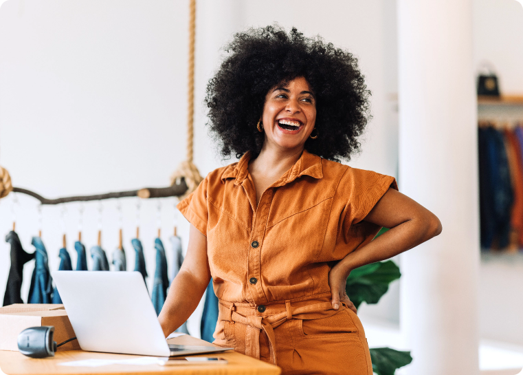 Business owner standing at a retail desk with a computer and box and a rack of clothing behind her