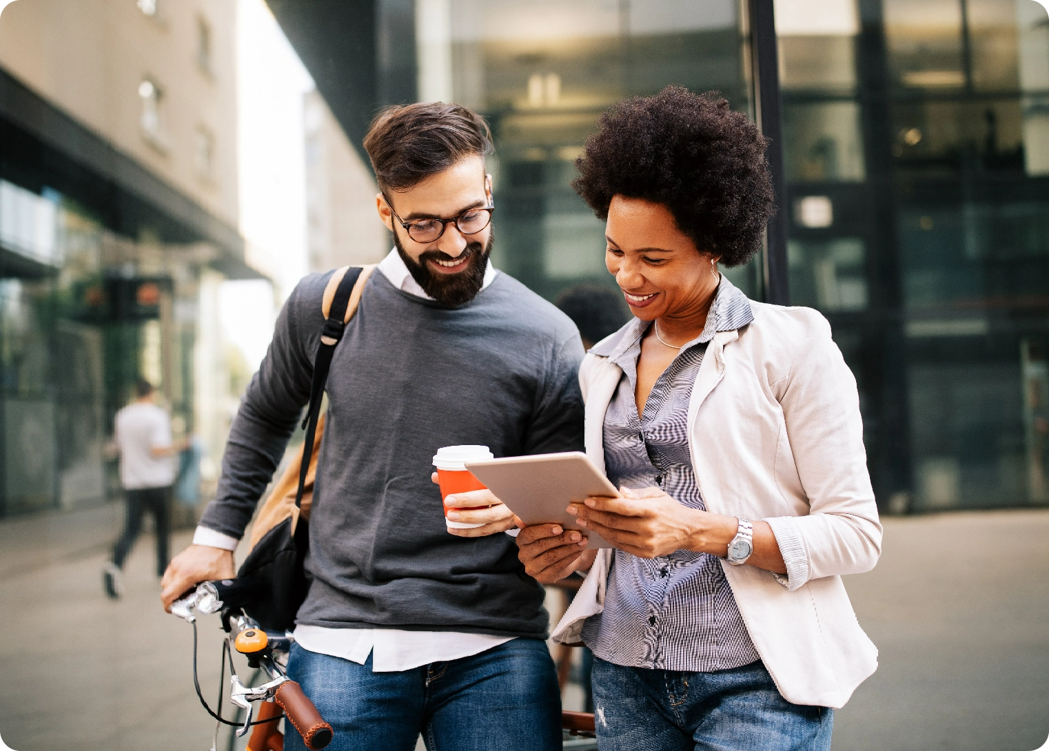 A man holding a leaning bike while hold a coffee cup and a woman standing next to him looking at a tablet and smiling