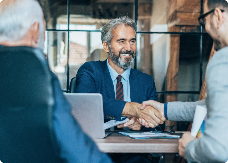 Two men in business attire shaking hands at a desk smiling at each other