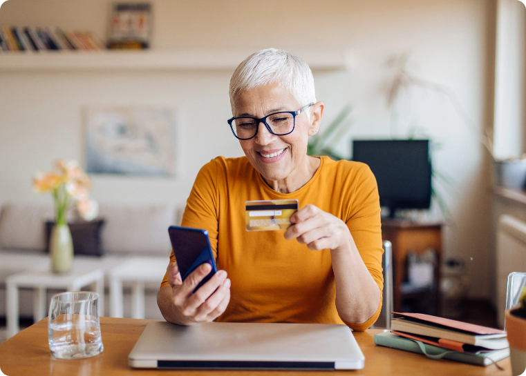 Woman sitting at a home desk looking at a mobile phone, holding a credit card and smiling