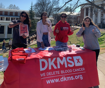 Four University of Virginia students smile while standing behind a DKMS registration table outside. Four University of Virginia students smile while standing behind a DKMS registration table outside.