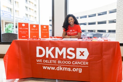 DKMS volunteer smiling while sitting at a table with a DKMS tablecloth, DKMS registration packets, giveaways, and signs. 