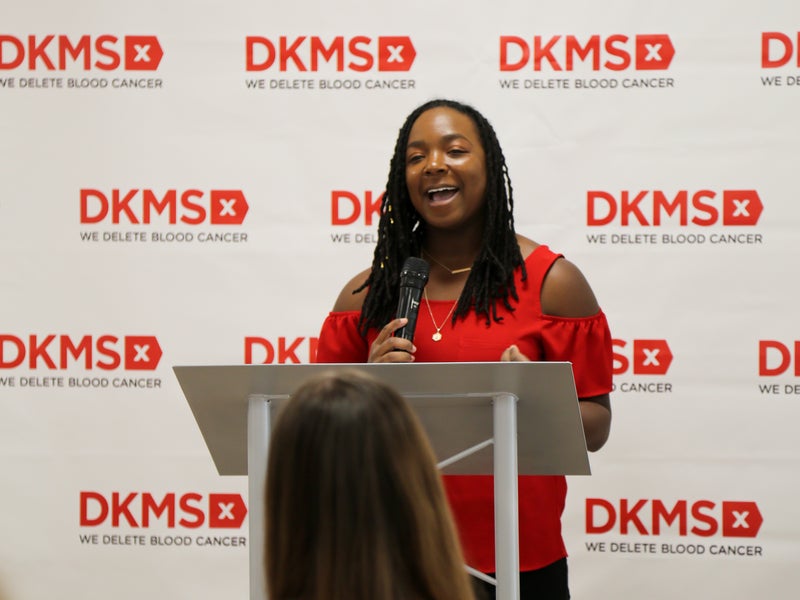DKMS Stem Cell Donor, Jasmine Deberry Thompson, standing at the podium giving a speech in front of a DKMS banner. 