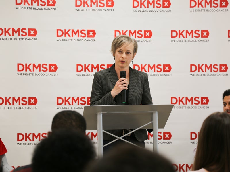 Stem cell transplant recipient, Kristi Martin, standing at the podium speaking in front of a DKMS banner 