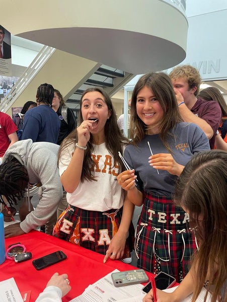 Two high school students wearing University of Texas shirts smile while doing a cheek swab to register for DKMS