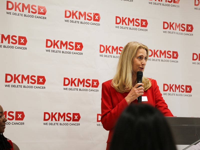 Dr. Elke Neujahr, DKMS Group Global CEO, stands in front of the podium speaking with the DKMS banner in the background.