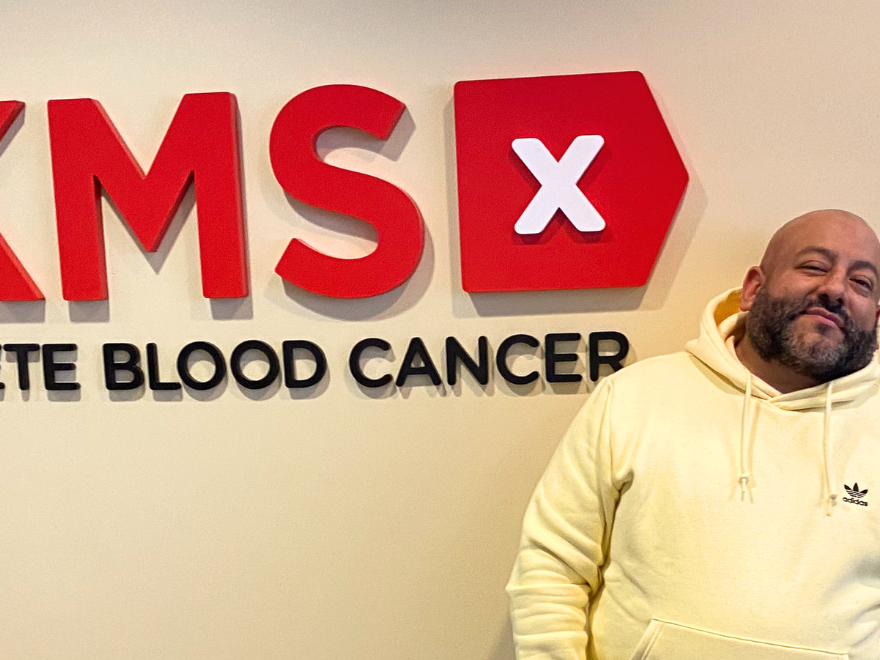 Pretty Lou wearing a yellow sweatshirt stands to the right of the DKMS logo in the NYC office Pretty Lou wearing a yellow sweatshirt stands to the right of the DKMS logo in the NYC office