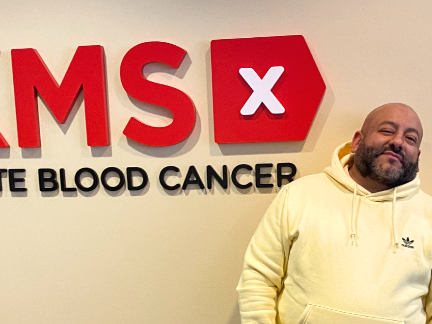 Pretty Lou wearing a yellow sweatshirt stands to the right of the DKMS logo in the NYC office Pretty Lou wearing a yellow sweatshirt stands to the right of the DKMS logo in the NYC office