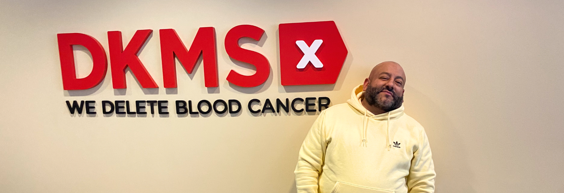 Pretty Lou wearing a yellow sweatshirt stands to the right of the DKMS logo in the NYC office
