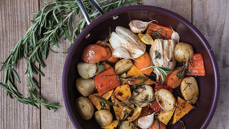 A purple non-stick frying pan with vegetables in on a wooden table with a bunch of rosemary.