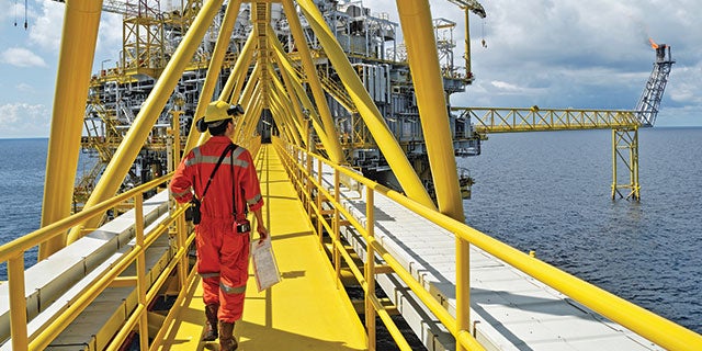 Ingegnere che indossa una tuta da lavoro arancione e un elmetto giallo che cammina su un ponte dipinto di giallo su un impianto energetico offshore.