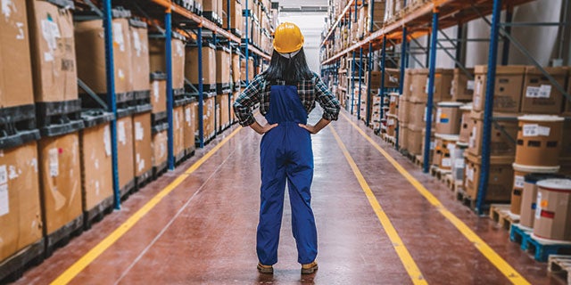 Woman wearing a yellow hardhat in the middle of a warehouse with shelves containing inventory.