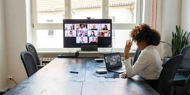 Woman sat at a meeting table waving at laptop screen to colleagues on video conferencing software who are working remotely.
