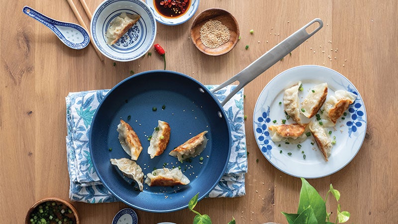 Fried gyoza in blue PPG/Whitford nonstick frying pan on a wooden table with kitchenware containing ingredients