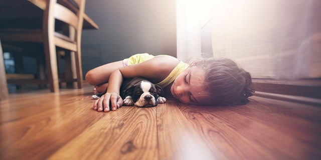 Una niña con su perro tumbados en un suelo de madera en casa. El aspecto del suelo se conserva con los revestimientos de madera de alto rendimiento de PPG.