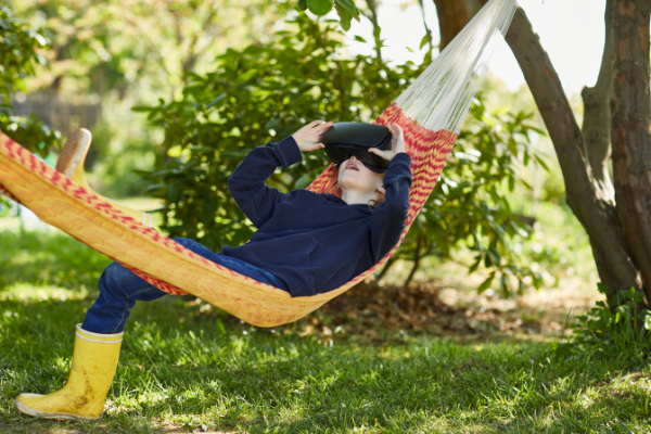 Young boy lying in a hammock in a garden as he looks through virtual reality glasses.
