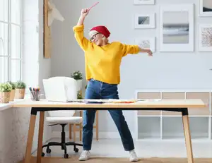 Frau im gelben Pulli tanzt vor einem Schreibtisch.