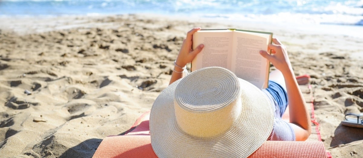 Person mit Hut am Strand, liest ein Buch.