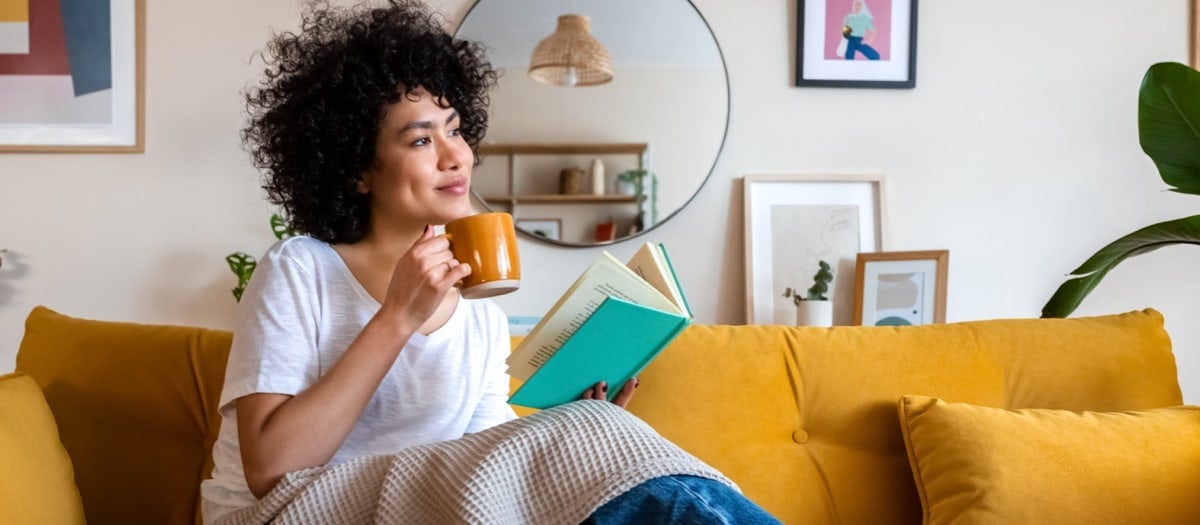 Frau sitz mit Tasse in der Hand auf einer Couch und liest.