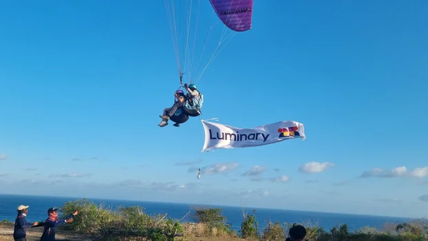 two people paragliding on a sunny day with the Luminary logo on a flag trailing behind them