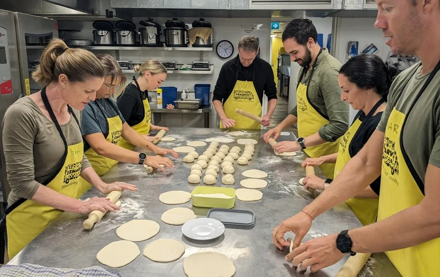 The Luminary team in yellow aprons around the kitchen table at OzHarvest, rolling dough