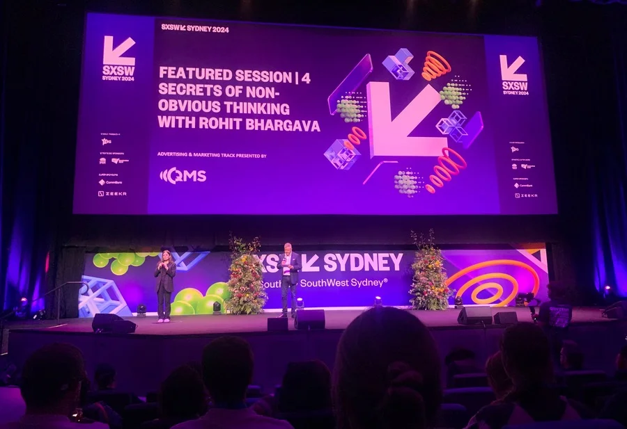 A purple screen with the SXSW logo on stage with 2 men speaking, infront of many seated people