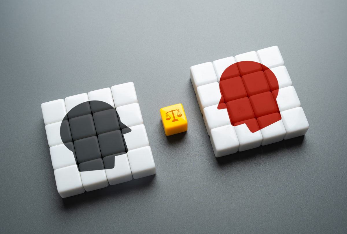 Two opposing heads, one black and one red, face away from one another. A yellow cube with an icon of balanced scales is placed in between