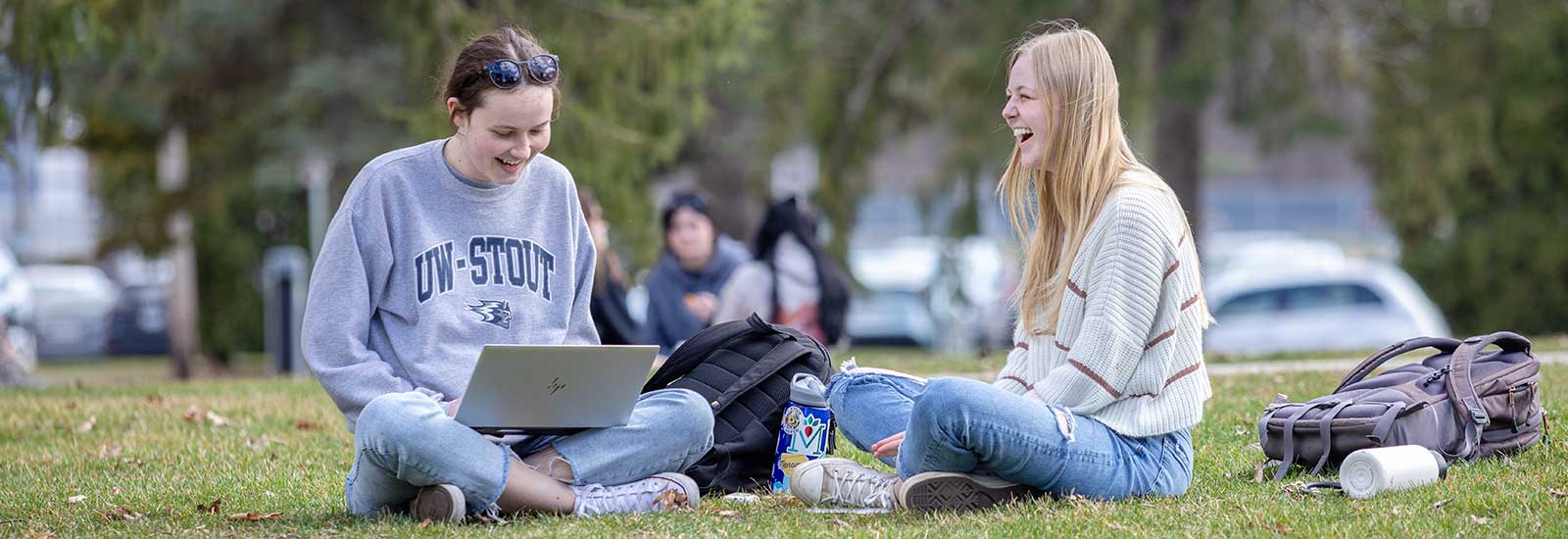 Two students sitting and laughing on grass with a laptop