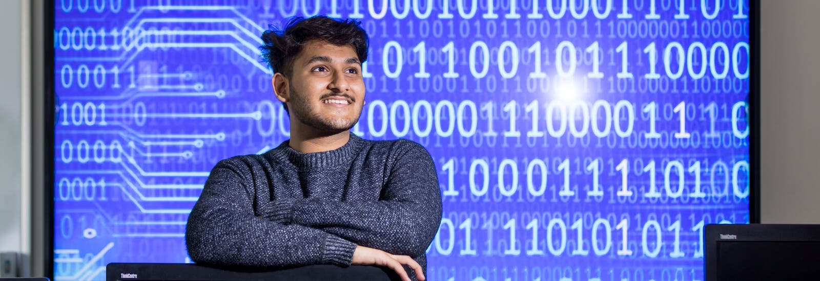 A student leaning on a computer, smiling, in front of screen