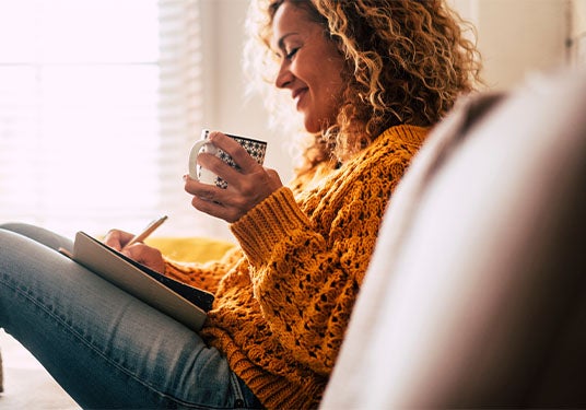 Woman happily writing in a journal on the couch with a coffee mug