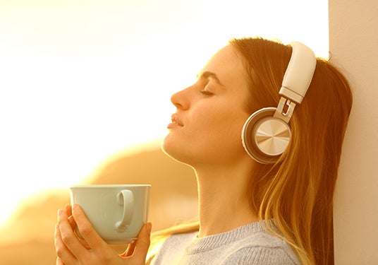 Woman drinking her coffee in the sun with headphones on and eyes closed