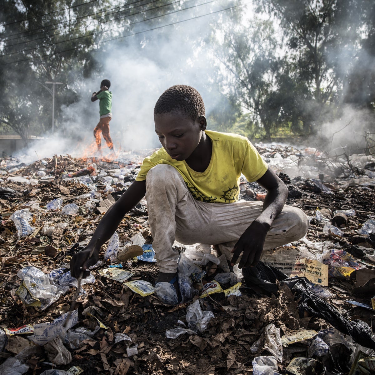 Teenager wearing a yellow shirt digs through burning rubbish dump for saleable items.