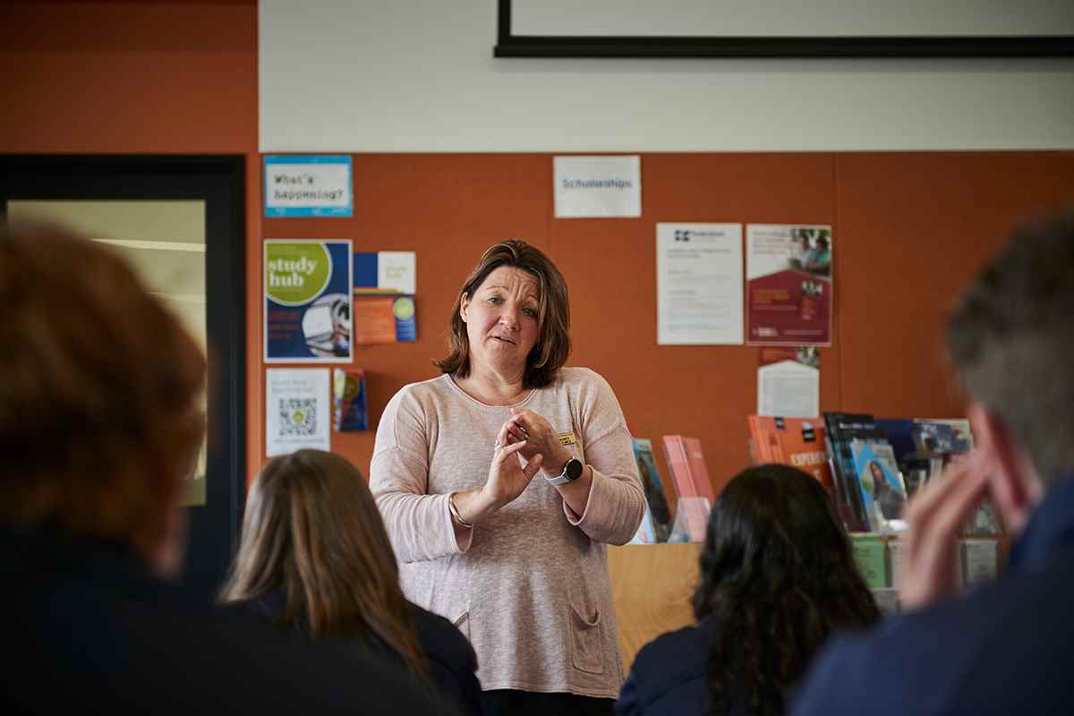 A teacher, facing the camera, stands in front of a class. An orange wall with posters and textbooks is in the background.