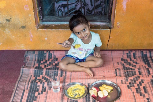 A young girl is seating on a carpet. She is showing a spoonful of food to the camera.