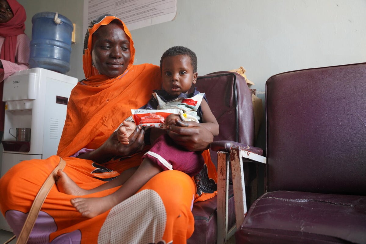 A child holding a food sachet, sitting with his mother on a chair. 