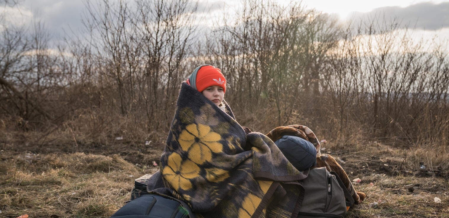 A young girl with blankets wrapped around her, sitting on luggage on the side of a road. 