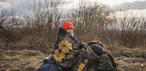 A young girl with blankets wrapped around her, sitting on luggage on the side of a road. 