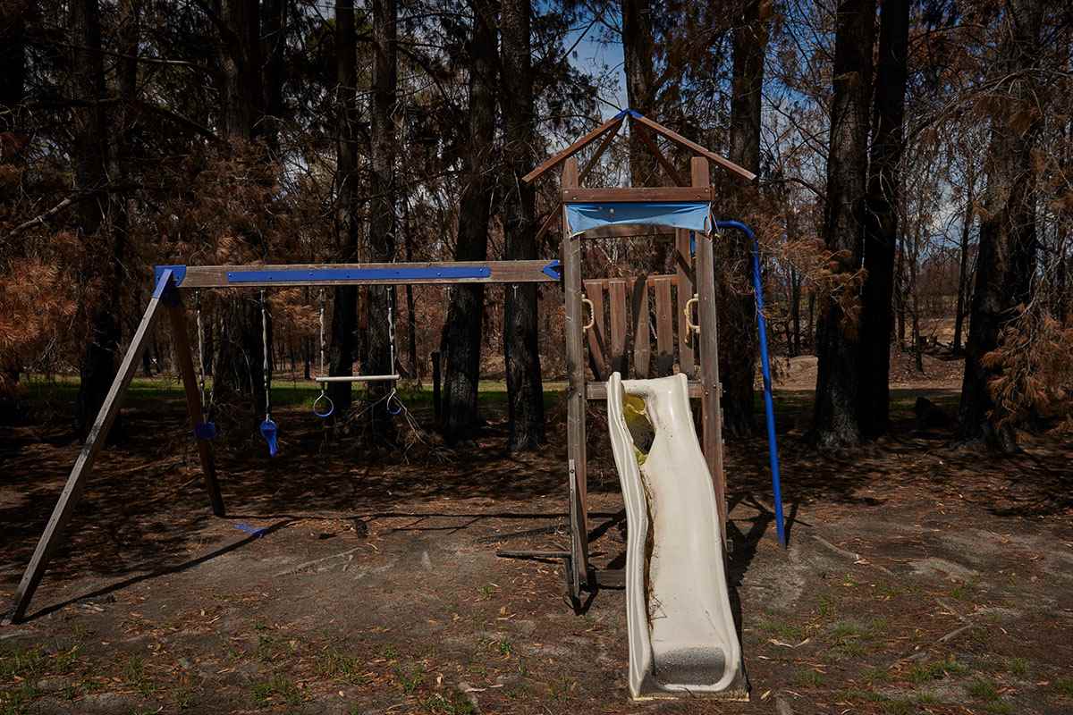 A set of play equipment surrounded by trees, with swings on the left and a slide on the right. The equipment is blackened and melted from the effects of fire.