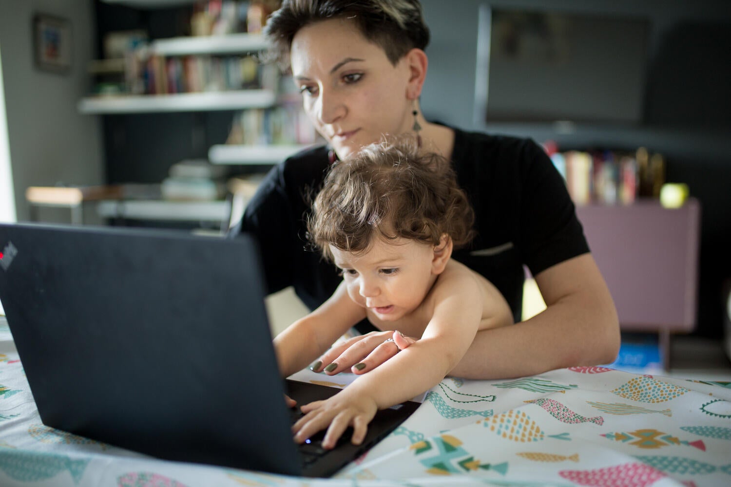 A woman is working on her laptop computer with a toddler on her lap. The toddles is aiming to use the computer's keyboard.