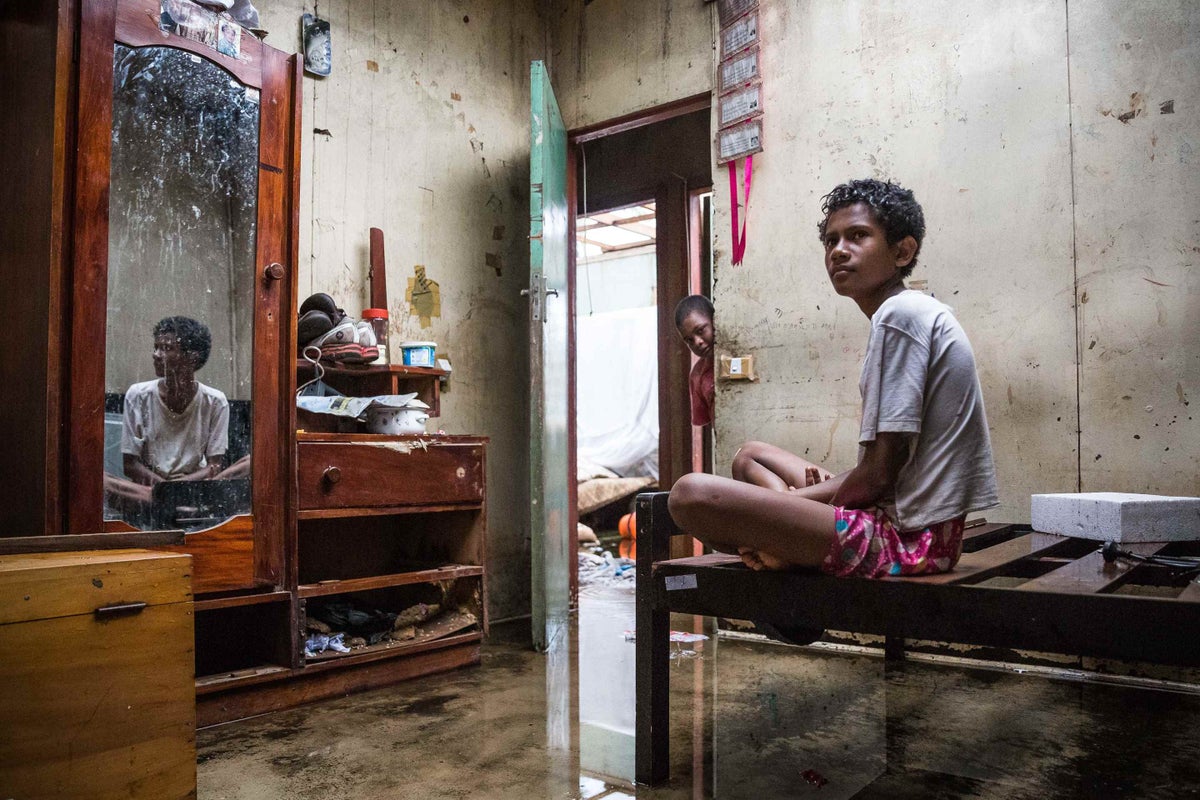 Makereta sitting on a wooden bed frame in what used to be her room. The roof is missing and sunlight streams through; she runs her feet through the water that covers the floor of her entire house.