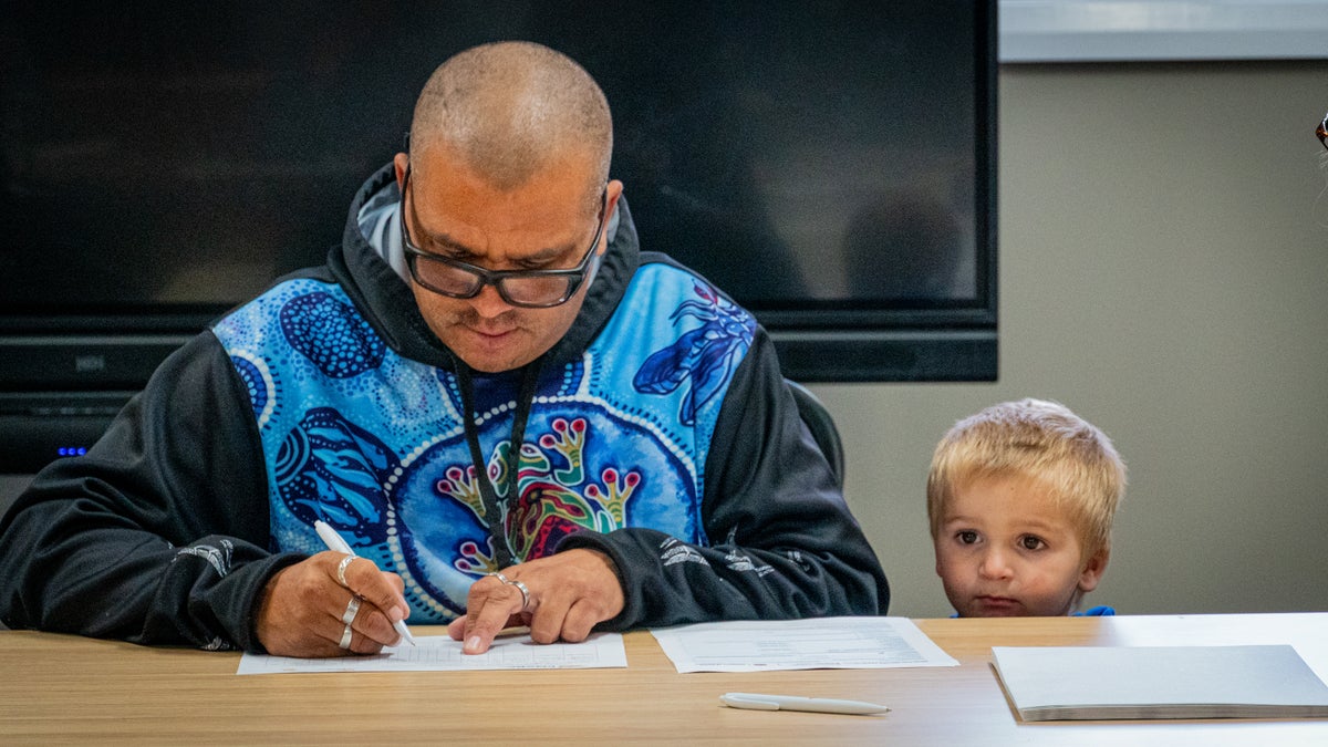 A dad signing a piece of paper on a table with his young son next to him. 