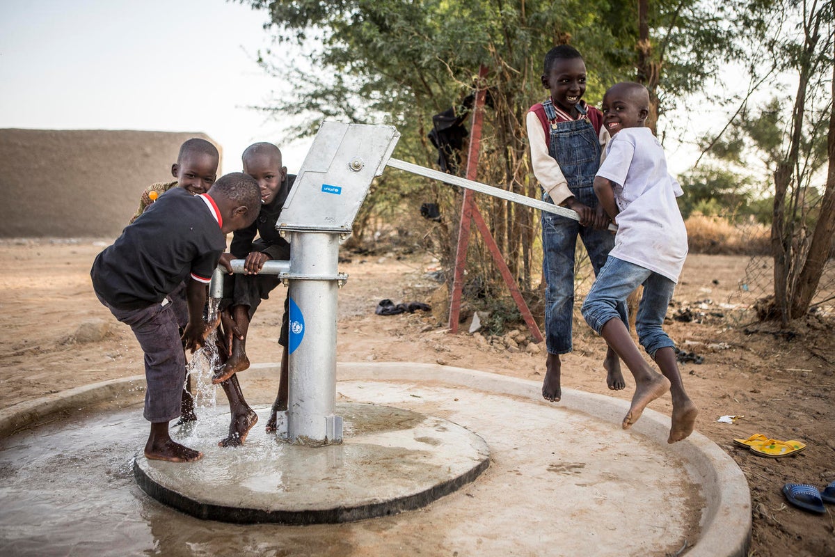 Two boys are actioning a manual water pump. Other three boys are on the tap side getting wet with the water that comes out of the tap. They all look happy.