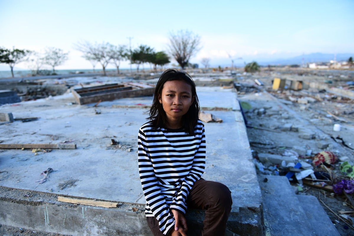 A young girl looks at the camera. The background is a destroyed area, there's only foundations for what used to be houses.