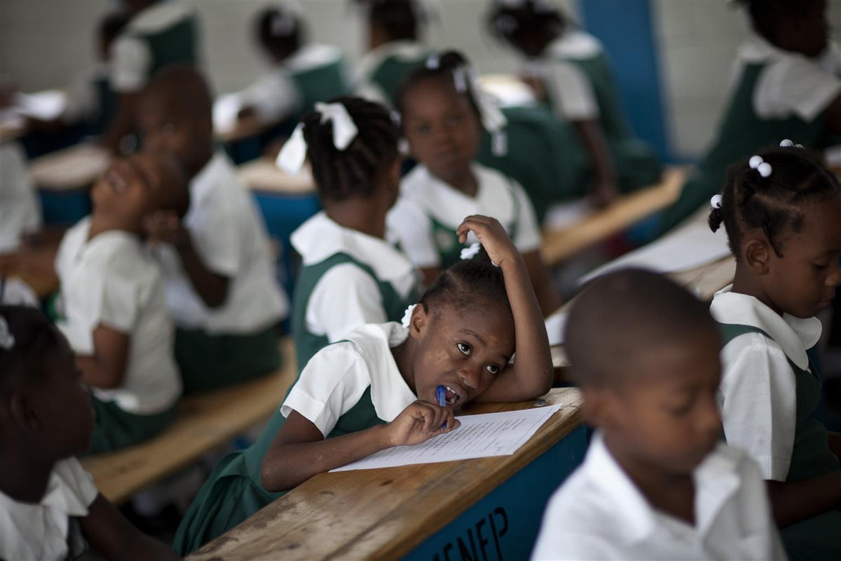 An image of a classroom with children. The image focuses on a girl that is looking at the teacher and writing on her notebook.