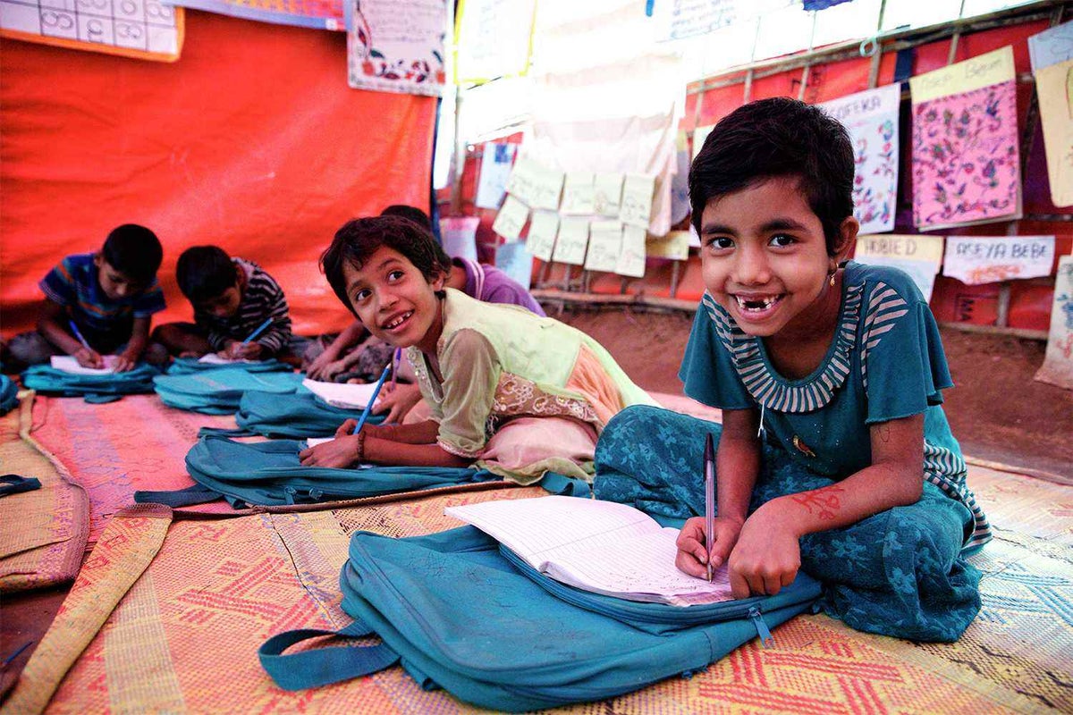 Children sitting on floor mats writing in notebooks. The closest child is smiling at the camera.