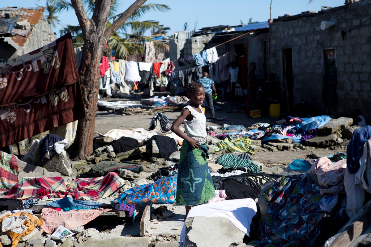 A girl walks through a damaged street. There's rugs and rubbish all over.